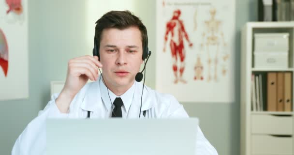 Mladý bělošský lékař ve sluchátkách sedí u stolu ve skříni. Doktorské videochatování na notebooku a konzultace s pacientem přes webkameru. Medik s videochat a konzultace on-line. — Stock video