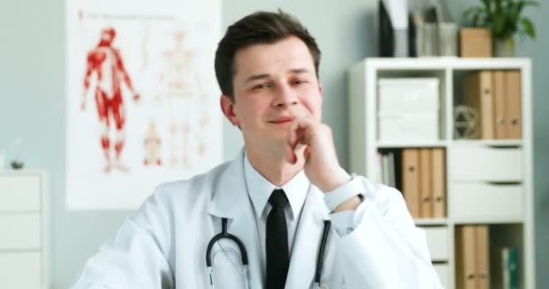 Portrét pohledného mladého bělocha zdravotníka v bílých šatech se stetoskopem sedícím ve skříni během pracovního dne. Dobře vypadající veselý mužský lékař otáčení tváří v tvář fotoaparátu a usmívání. — Stock video