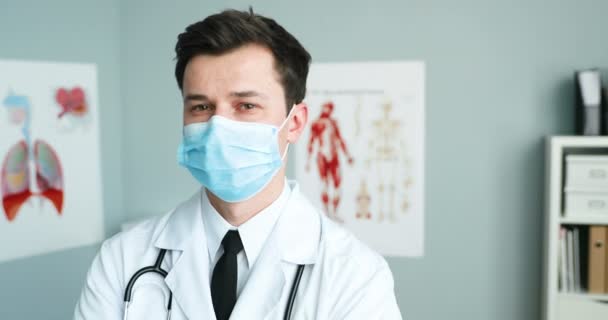 Προσωπογραφία ενός όμορφου νεαρού Καυκάσου γιατρού με λευκό φόρεμα, ιατρική μάσκα και στηθοσκόπιο να στέκεται στο ντουλάπι και να κοιτάζει την κάμερα. Αρσενικός γιατρός κατά τη διάρκεια της πανδημίας του κορωνοϊού. Έννοια προστασίας. — Αρχείο Βίντεο