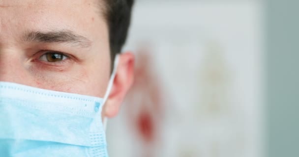 Κοντινό πλάνο του μισού προσώπου του νεαρού θλιμμένου Καυκάσου γιατρού με μπλε ιατρική μάσκα που κοιτάζει την κάμερα. Άντρας γιατρός στην αναπνευστική προστασία στο ντουλάπι. Πανδημία του Coronavirus. — Αρχείο Βίντεο