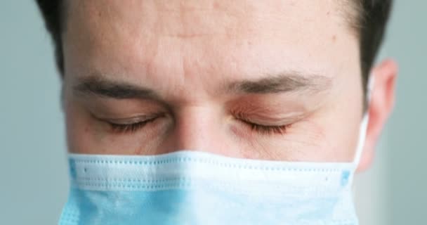 Close-up da face de branco jovem médico do sexo masculino em azul máscara médica olhando para baixo e subindo os olhos para a câmera. Doutor em desespero. pandemia de coronavírus. Médico de protecção respiratória . — Vídeo de Stock