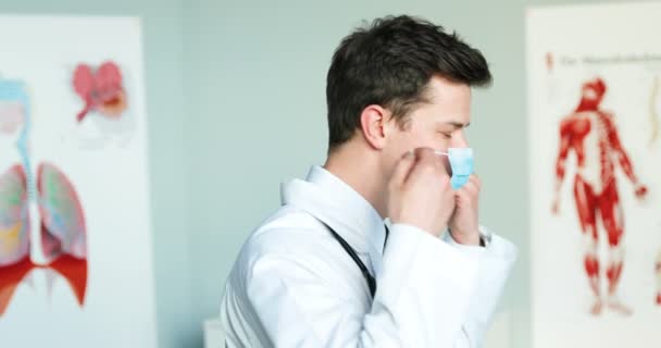 Giovane medico caucasico bello maschio in abito bianco assumendo maschera medica blu. Protezione respiratoria durante l'epidemia di coronavirus per il medico. Uomo medico girando il viso e guardando la fotocamera. — Video Stock