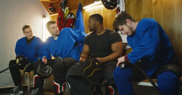 Joueurs de hockey masculins de races mixtes assis dans la cabine d'essayage et parlant après le match. Des sportifs multiethniques discutent et vérifient leur équipement dans le vestiaire pendant la pause. — Video