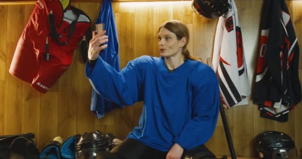 Bastante joven jugador de hockey femenino caucásico en uniforme azul sentado en el vestidor y tomando fotos selfie con la cámara del teléfono inteligente. Hermosa deportista posando y sonriendo al teléfono en el vestuario . — Vídeos de Stock