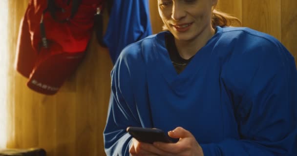 Close-up van vrolijke en zweterige jonge blanke hockeyspeelster die op de bank zit in de kleedkamer en sms 't op de smartphone. Sportvrouw in kleedkamer afluisteren en scrollen op de telefoon. — Stockvideo