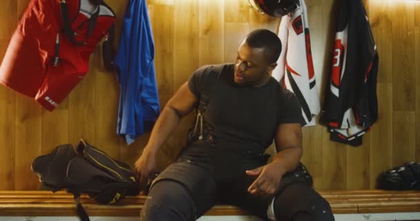 Retrato de un joven guapo y fuerte jugador de hockey afroamericano sentado en el banco en el vestidor, quitándose el uniforme y sonriendo alegremente a la cámara. Después del juego en el vestuario . — Vídeos de Stock