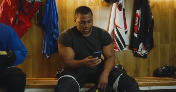 Afroamerikanska unga manliga hockeyspelare sitter på bänken i omklädningsrum och sms: ar meddelande på smartphone. Snygg idrottsman knacka och rulla på mobiltelefon i omklädningsrummet. — Stockvideo