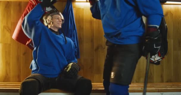 Veselý mladý kavkazský mužský hokejista přichází do šatny za zápasem, sedí na lavičce a svléká uniformy. Potící se šťastní sportovci mluví a usmívají se v šatně. — Stock video