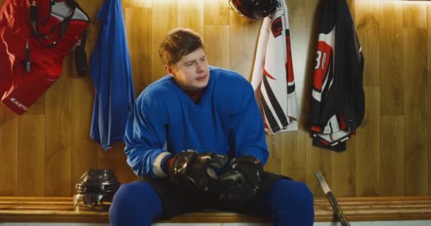 Portrait d'un joueur de hockey masculin caucasien en colère en uniforme bleu assis sur un banc dans un vestiaire et regardant tristement la caméra. Sportif disculpé dans le vestiaire après avoir perdu. — Video