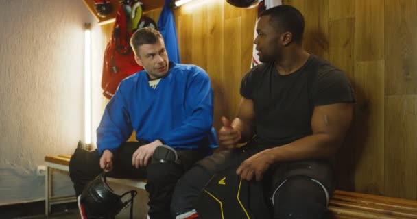 Jugadores de hockey masculino de carreras mixtas sentados en el banco en el vestidor y hablando mientras se preparan para el juego. Deportistas hablando en vestuario y vistiendo. Amigos multiétnicos animándose antes del juego . — Vídeos de Stock