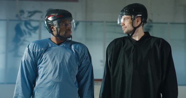 Üniformalı ve kasklı çok ırklı erkek hokey oyuncularının birbirlerine bakıp buz arenasında kameraya gülümseyişlerinin portresi. Melez sporcular ayakta ve maça hazırlanıyor.. — Stok video