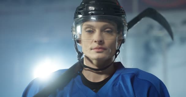 Portrét záběru vážně krásné mladé bělošky hokejistky v helmě a s klubem dívá na kameru na ledové aréně. Zblízka tvář atraktivní sportovkyně v casque s klackem.