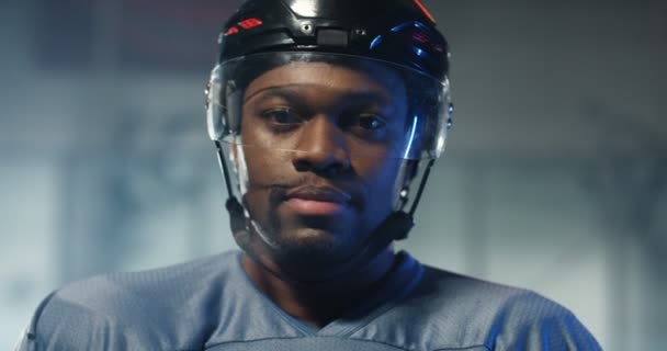 Крупный план красивого молодого афроамериканского хоккеиста в шлеме, поворачивающегося лицом и смотрящего в камеру с проницательными глазами на ледяной арене. Портрет спортсмена в каске. — стоковое видео