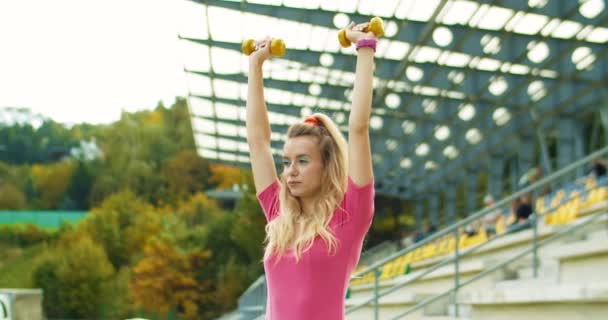 Porträt einer schönen kaukasischen Sportlerin, die im Freien mit Kurzhanteln über dem Kopf Pressearbeit macht. Lustige fröhliche hübsche Mädchen im Retro-Stil im rosafarbenen Body beim Training. Retro-Konzept — Stockvideo