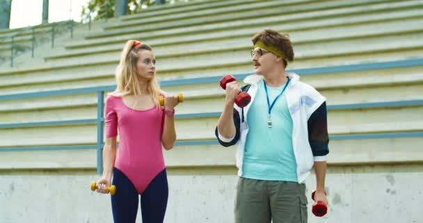 Vintage handsome funny fitness trainer showing to pretty girl in pink bodysuit exercises on biceps on street. Male and female athletes working out with dumbbells outdoors near stairs. 70s concept — Stock Video