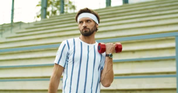 Kaukasischer Retro-Mann mit Stirnband und lustigem Gesichtsausdruck beim Training auf der Straße. Porträt eines gut aussehenden Sportlers im Retro-Stil, der mit roten Kurzhanteln im Freien Übungen auf dem Bizeps macht. Vintage-Konzept — Stockvideo