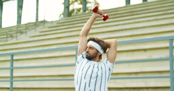 Portrét pohledného retro sportovce, jak cvičí na tricepsech s červeným činkou a dívá se ven. Bělošský vtipný veterán v čelence, co cvičí na ulici. Koncept zdravého života — Stock video