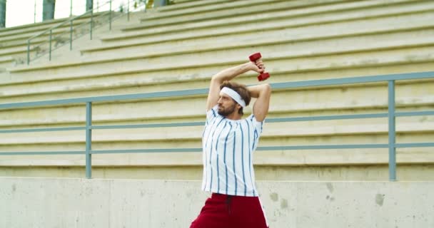 Blanke grappige vintage fitnessman in hoofdband die op straat traint. Stomme knappe retro sporter doet oefeningen op triceps met rode halter en kijkt weg buiten. Sportconcept — Stockvideo