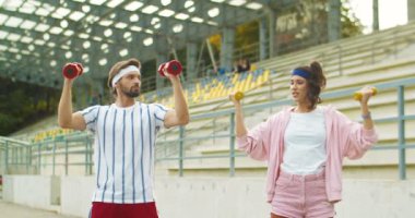 Eski moda yakışıklı fitness eğitmeni, stadyumdaki klasik kız egzersizlerini gösteriyor. Erkek ve bayan komik sporcular dışarıda küçük dambıllarla antrenman yapıyorlar. Spor yaşam tarzı konsepti