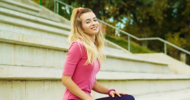 Portret van een prachtig vrolijk vintage meisje zittend op tribune en glimlachend naar de camera. Vrolijke mooie blanke jonge retro stijl vrouw in paarse legging en roze bodysuit outdoor. 70s-concept — Stockvideo