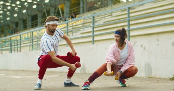 Vintage przystojny zabawny mężczyzna i ładna dziewczyna robi ćwiczenia na bicepsach na ulicy. Mężczyzna i kobieta głupio wyglądający sportowcy w stylu retro ćwiczą z hantlami na świeżym powietrzu w pobliżu schodów. Koncepcja lat 70. — Wideo stockowe