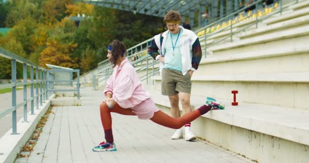 Instrutor atlético masculino caucasiano engraçado olhando para menina vintage bonita que está fazendo lunges de perna única em escadas ao ar livre. Bonita esportista estilo retro trabalhando com treinador. Conceito de desporto — Vídeo de Stock