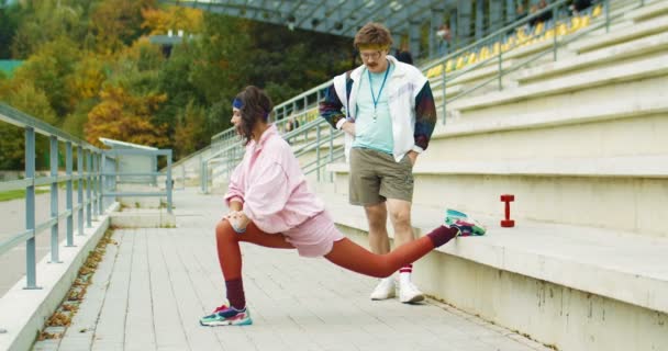 Grappige blanke mannelijke atletische instructeur op zoek naar mooie vintage meisje die doet single-leg lunges op trappen buiten. Prachtige retro stijl sportvrouw aan het trainen met de coach. Sportconcept — Stockvideo