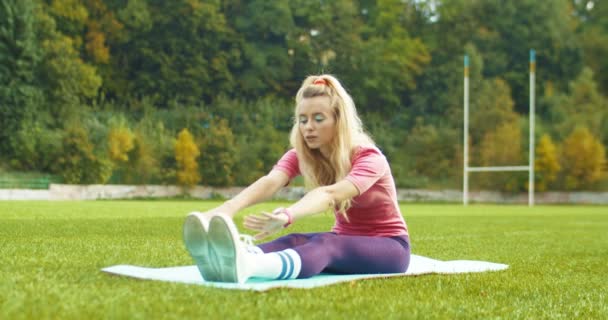 Vacker vintage blond idrottsman i rosa body suit värma upp och göra övningar medan du sitter. Ganska vit ung retro-stil sportskvinna sträcker muskler på yogamattan utomhus. Begreppet ändamålsenlighet — Stockvideo