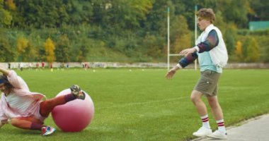 Komik, mutlu, beyaz, güzel, klasik bayan atlet dışarıda fitness topuna atlayıp yere düşüyor. Bıyıklı erkek fitness hocası ona yardım ediyor. Spor yaşam tarzı. Retro kavramı