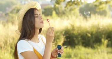 Beyaz tenli, şapkalı, esmer bir kız yazın doğada sabun köpüğü üflüyor. Genç ve güzel bir bayan güneşli bir günde dışarıda oynuyor..