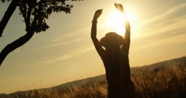 Koyu renk siluet şapkalı genç bir kadın yazın güneşinde doğada dikiliyor. Arka planda elleri havada ve arka planda güneşi olan bir kız var. Tatil kavramı.
