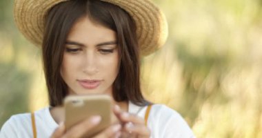 Güneşli bir günde, şapkalı, akıllı telefonları dinleyen ve mesaj atan hoş, beyaz bir kız. Yazın doğada cep telefonunda yazı yazan ve kaydıran güzel esmer kadın..