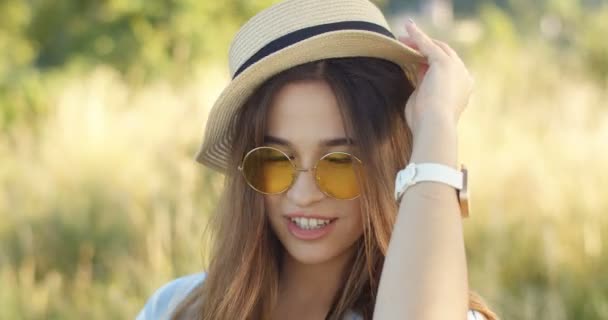 Närbild av vackra unga kaukasiska flicka med långt mörkt hår och i solglasögon ler mot kameran och tar på sig hatt. Porträtt av vacker kvinna i naturen på solig dag. — Stockvideo