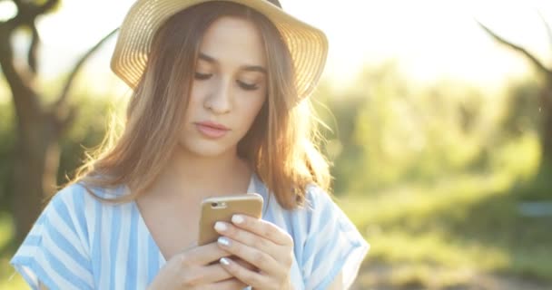 Şapkalı güzel beyaz bir kız. Akıllı telefonları dinliyor ve yazın açık havada mesaj yolluyor. Güneşli bir günde doğada cep telefonunda yazan ve kaydıran güzel esmer kadın.. — Stok video