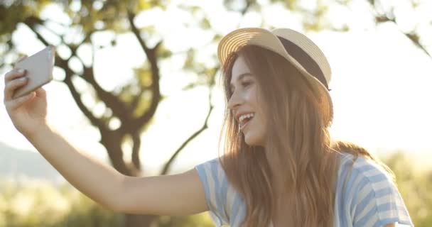 Mooi blank jong meisje met donker haar en een hoed lachend en poserend voor smartphone camera op zonnige dag in de natuur. vrij vrolijk brunette vrouw het nemen van foto 's met telefoon in de zomer. — Stockvideo