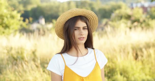 Portret van een mooie jonge brunette blanke vrouw met hoed tanding in het veld en kijkend naar de camera. Charmant meisje met donker haar in de natuur plaats in de zomer. Buiten. — Stockvideo