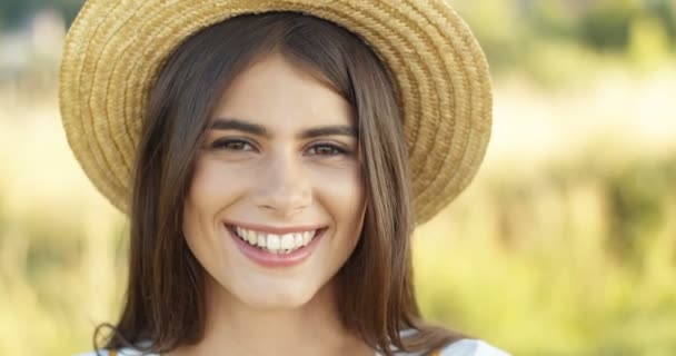 Close-up van mooie vrolijke Kaukasische brunette meisje met hoed glimlachen gelukkig om camera outdoor in de zomer. Portret van charmante jonge vrouw in goed humeur met glimlach op de natuur. — Stockvideo