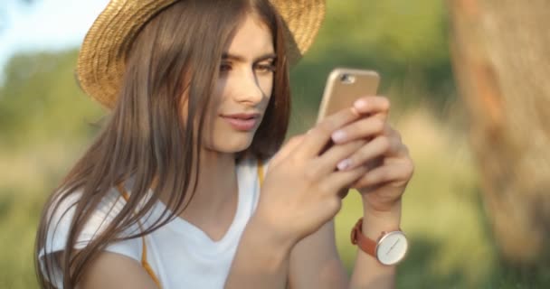 Vacker ung kaukasisk kvinna i hatt och med långt mörkt hår sitter i naturen på solig sommardag och sms:ar samtidigt som du avlyssnar telefonen. Söt flicka chatta och bläddra på smartphone. — Stockvideo