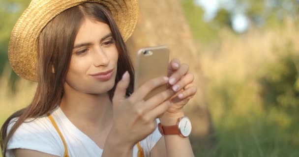 Närbild av vackra unga kaukasiska kvinna i hatt och med långt mörkt hår sitter i naturen på sommaren dag och sms: a meddelande medan du avlyssnar på telefon. Söt flicka chatta och bläddra på smartphone. — Stockvideo