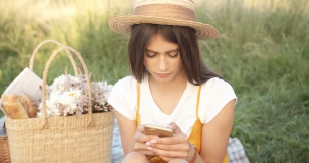 Vacker ung kaukasisk kvinna i hatt och med långt mörkt hår sitter i naturen på picknick och SMS samtidigt knacka på telefonen. Söt flicka chatta och bläddra på smartphone. — Stockvideo