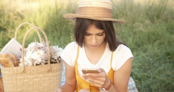 Vacker ung vit kvinna i hatt med mörkt hår sitter på naturen på picknick med persika och sms: a, knacka på telefonen. Söt flicka chatta och rulla på smartphone med frukt i handen. — Stockvideo