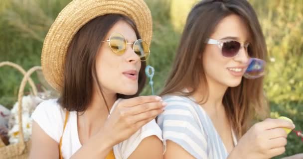 Närbild av ganska glada flickor, bästa vänner i hatt annons solglasögon sitter tillbaka på grönt gräs på sommaren med korg och blåser såpbubblor. Vackra kvinnor som leker, har kul på picknick. — Stockvideo