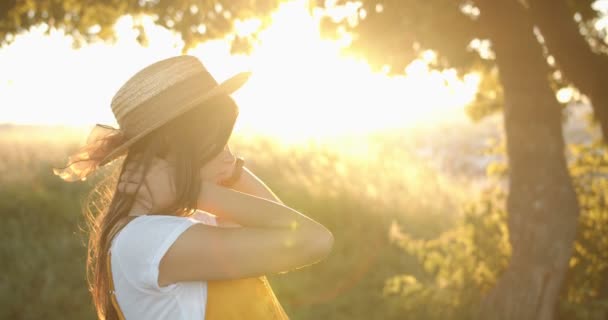 Портрет молодой красивой белой девушки в шляпе, позирующей перед камерой и радостно улыбающейся в камеру на закате солнца на природе. Красивая женщина с улыбкой, стоящая на дереве в поле с сияющим солнцем позади — стоковое видео