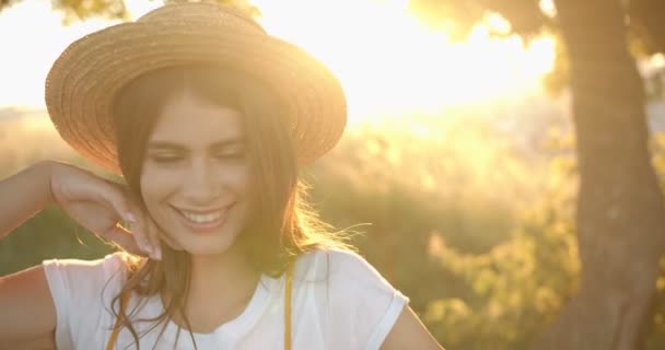 Κοντινό πλάνο του νεαρού όμορφου καυκάσιου κοριτσιού με καπέλο που ποζάρει στην κάμερα και χαμογελά χαρούμενα στην κάμερα στο ηλιοβασίλεμα στη φύση. Πορτρέτο της όμορφης γυναίκας με χαμόγελο στέκεται στο δέντρο στον τομέα με ήλιο λάμπει — Αρχείο Βίντεο