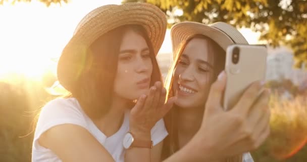 Två vita unga glada flickor i hattar tar selfie foton med smartphone kamera i solljus. Glada vackra bästa vänner ler för att ringa när du gör foto på naturen. Fotografering utomhus. — Stockvideo