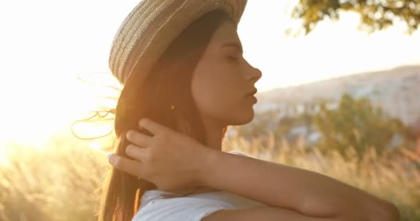 Close-up van de jonge Kaukasische mooie meisje met hoed met lange brunette haar draaien gezicht naar camera en vrolijk glimlachen bij zonsondergang op de natuur. Portret van vrij charmante vrouw in het veld in de zomer. — Stockvideo