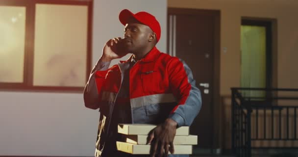 Піца афроамериканець у червоній уніформі і кашкет стоїть біля будинку з піцерійними коробками і телефонує ввечері. На вулиці. — стокове відео