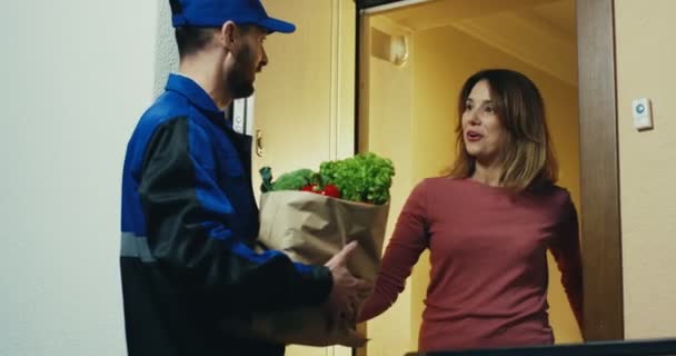 Кавказький чоловік у синій формі з супермаркету дає пачку з овочами привабливій жінці біля її дверей. У приміщенні. — стокове відео