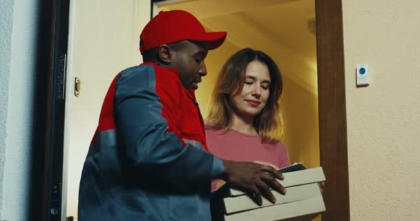 Afro-Amerikaanse jongeman in uniform en pet die pizza aflevert aan de blanke vrouw. Binnenkant. — Stockvideo