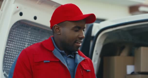 Крупный план афроамериканского молодого курьера в красной форме и кепке, стоящих на улице у белого фургона и подсчитывающих почтовые ящики. — стоковое видео